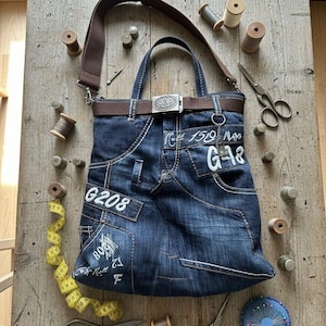 May include: A denim tote bag with a brown strap and belt, featuring white stitching and pockets. The bag has the text "G208" and "G-18" on it. Sewing supplies like scissors, spools of thread, and a measuring tape surround the bag.