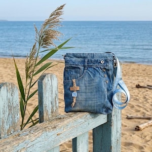 Könnte beinhalten: Eine Umhängetasche aus recycelten Jeans, mit einer Fronttasche, Knopfverschluss und einem hellblauen Riemen. Die Tasche ist mit einem kleinen Holzanhänger mit der Aufschrift "Too Cute Deluxe" verziert. Die Tasche wird auf einem verwitterten blauen Zaun mit Strand und Ozean im Hintergrund präsentiert.