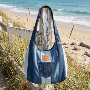 Könnte beinhalten: Jeans-Tote-Tasche mit Streifenmuster in Blau- und Weißtönen. Mit einer Vordertasche und einem Levi's-Markenaufnäher. Die Tasche hängt an einem verwitterten weißen Zaunpfosten, mit Strand und Meer im Hintergrund.
