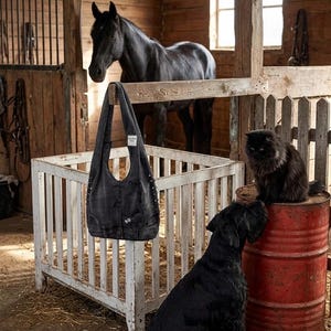May include: A black denim tote bag with silver stud accents hangs in a rustic barn setting. A black horse, a black cat, and a black dog are also present. The bag has pockets and a curved top.