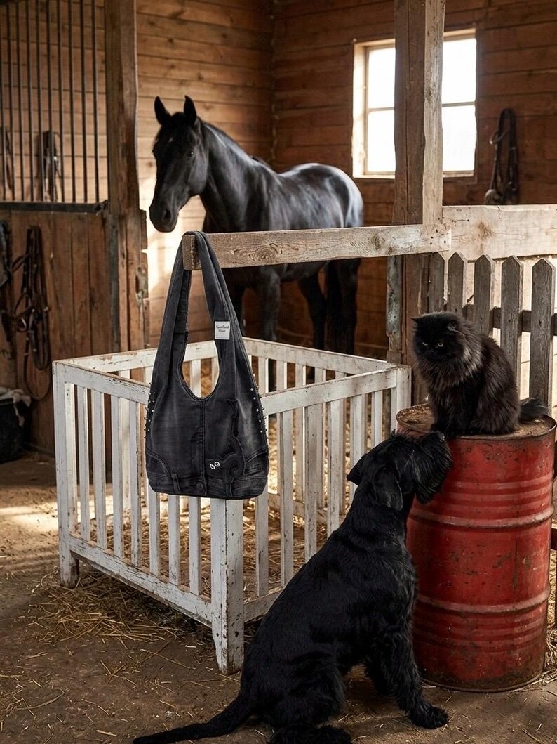 May include: A black denim tote bag with silver stud accents hangs in a rustic barn setting. A black horse, a black cat, and a black dog are also present. The bag has pockets and a curved top.