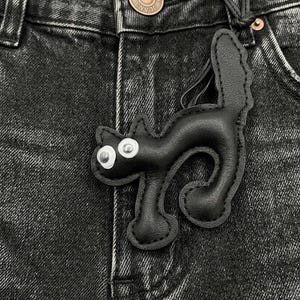 May include: A black leather cat-shaped keychain with white eyes and a stitched outline. The keychain is attached to a black leather loop, set against a backdrop of black denim jeans.