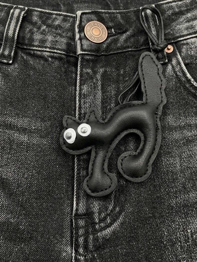 May include: A black leather cat-shaped keychain with white eyes and a stitched outline. The keychain is attached to a black leather loop, set against a backdrop of black denim jeans.
