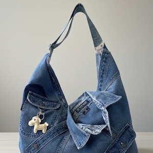May include: A light blue denim shoulder bag made from recycled jeans. The bag has a distinctive design with pockets and a denim strap. A small, cream-coloured horse keyring hangs from the front pocket. The bag is a light blue colour.