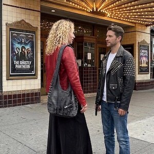 May include: A couple stands outside a vintage theater. The woman wears a red leather jacket, black skirt, and a gray denim shoulder bag. The man wears a black studded leather jacket, blue jeans, and black sneakers. A movie poster is visible.