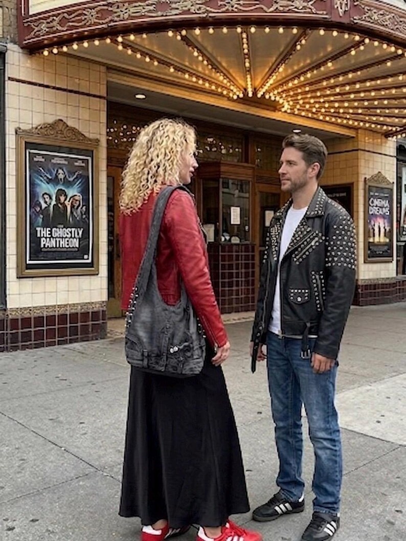 May include: A couple stands outside a vintage theater. The woman wears a red leather jacket, black skirt, and a gray denim shoulder bag. The man wears a black studded leather jacket, blue jeans, and black sneakers. A movie poster is visible.