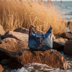 May include: A blue denim shoulder bag with brown leather straps and accents. The bag features the word "lions" embroidered in a light brown thread. The bag is sitting on a large rock near the water.
