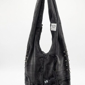 May include: A gray denim shoulder bag with silver-tone metal studs and a black cat-shaped embellishment. The bag has a zipper closure and a "Hand Made Denim" tag.