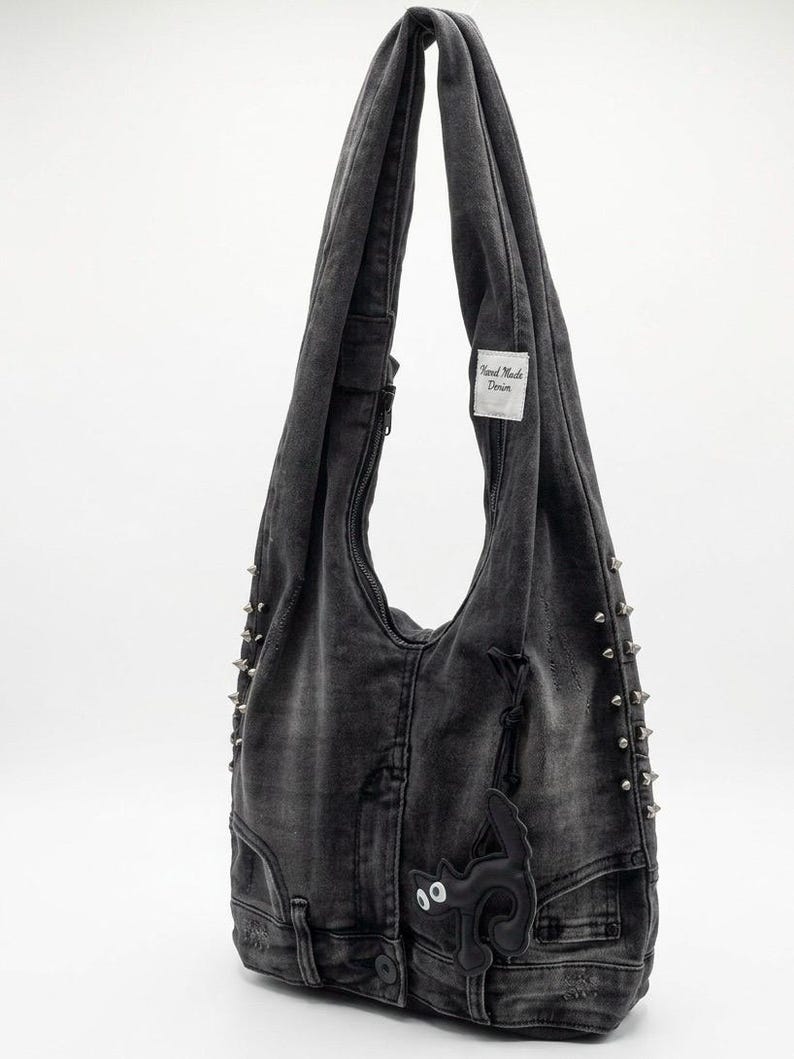 May include: A gray denim shoulder bag with silver-tone metal studs and a black cat-shaped embellishment. The bag has a zipper closure and a "Hand Made Denim" tag.