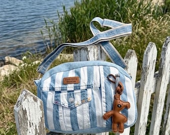 May include: A light blue and white striped denim crossbody bag featuring a front pocket with a button, a brown leather-like tag labelled Two Step Denim, and a brown dog-shaped charm attached to the side. It has a matching patterned shoulder strap.