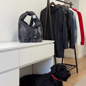 May include: A denim tote bag with silver studs and a white tag. The bag is made from recycled denim and has a unique, open design. A black dog with a red collar is lying on the floor near a white dresser and a clothing rack.