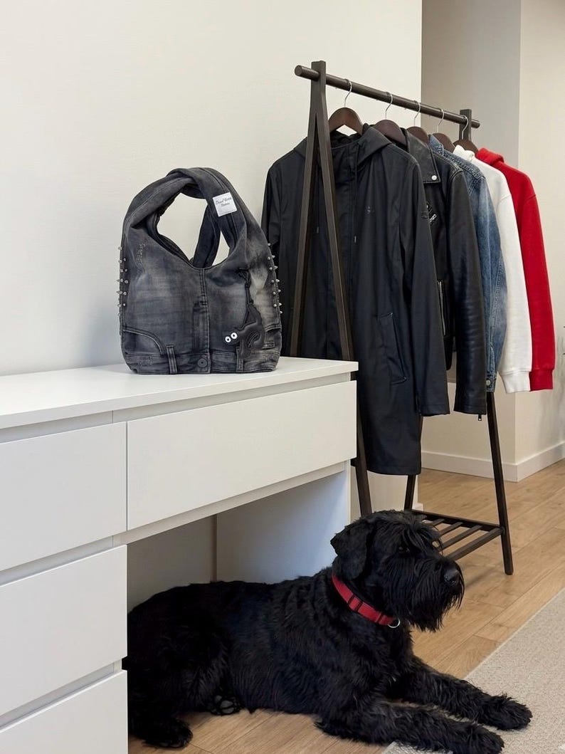 May include: A denim tote bag with silver studs and a white tag. The bag is made from recycled denim and has a unique, open design. A black dog with a red collar is lying on the floor near a white dresser and a clothing rack.