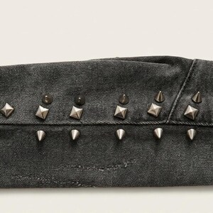 May include: Close-up of a section of dark gray denim fabric, embellished with silver-colored metal studs. The studs are a mix of cone and square shapes, adding a punk rock aesthetic to the garment.
