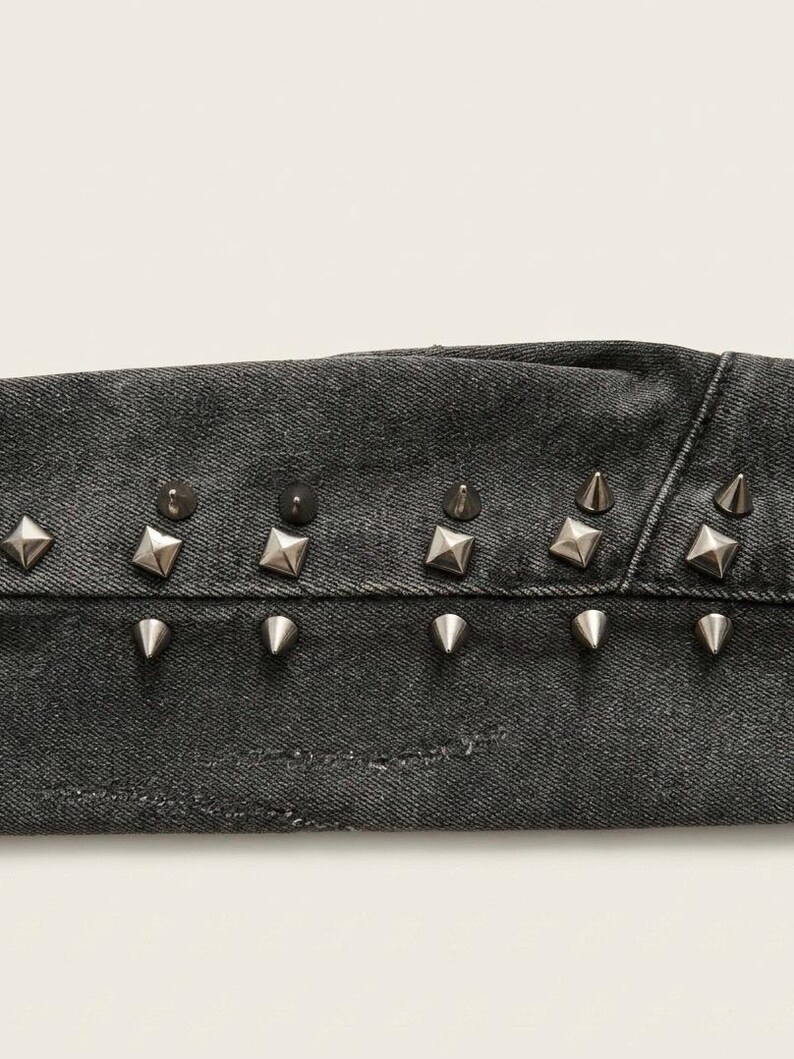 May include: Close-up of a section of dark gray denim fabric, embellished with silver-colored metal studs. The studs are a mix of cone and square shapes, adding a punk rock aesthetic to the garment.