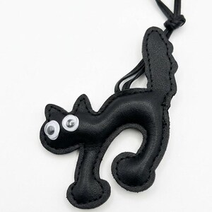 May include: Black leather cat-shaped keychain with googly eyes. The cat has a curved body, a tail, and a loop for attaching to keys or a bag. The keychain is made of stitched leather and has a simple, playful design.
