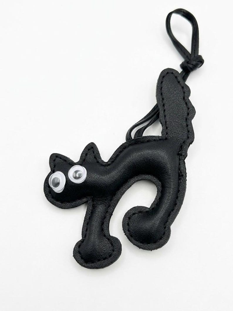 May include: Black leather cat-shaped keychain with googly eyes. The cat has a curved body, a tail, and a loop for attaching to keys or a bag. The keychain is made of stitched leather and has a simple, playful design.