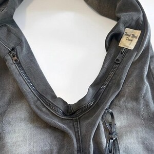 May include: Close-up of a gray denim bag with a zipper closure. The bag has a distressed look and a small tag that reads "Hand Work Denim". The bag is made of a durable material and has a unique design.