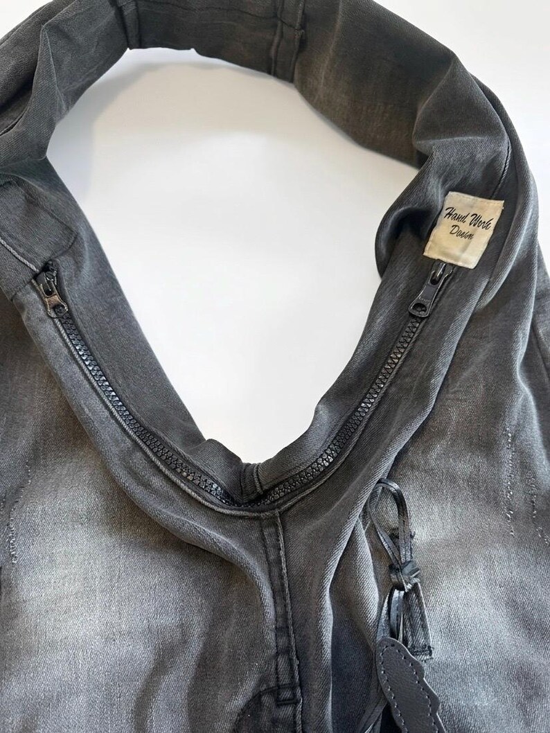 May include: Close-up of a gray denim bag with a zipper closure. The bag has a distressed look and a small tag that reads "Hand Work Denim". The bag is made of a durable material and has a unique design.