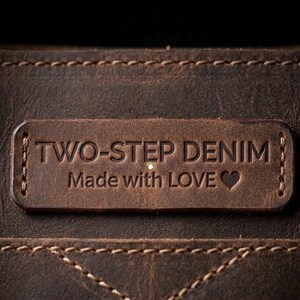 May include: Close-up of a brown leather tag with the embossed text "TWO-STEP DENIM" and "Made with LOVE" with a heart symbol. The tag is stitched onto a textured leather surface, showcasing a rustic aesthetic.