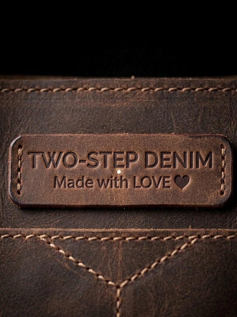 May include: Close-up of a brown leather tag with the embossed text "TWO-STEP DENIM" and "Made with LOVE" with a heart symbol. The tag is stitched onto a textured leather surface, showcasing a rustic aesthetic.