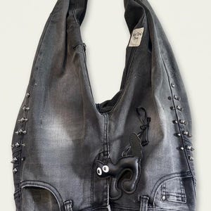 May include: Gray denim tote bag with silver-tone stud accents. Features a black leather cat applique and a tag that reads "Best Work Done". The bag has a button closure and a unique design.