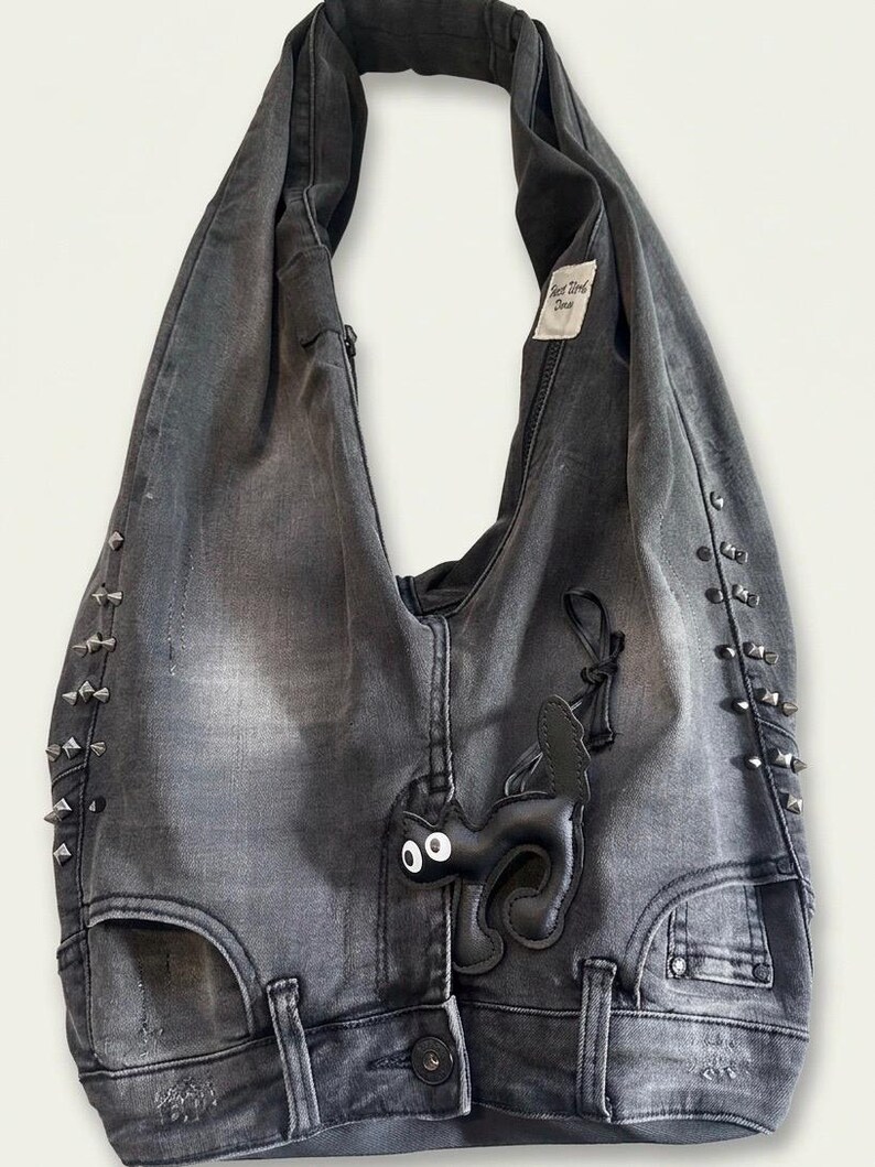 May include: Gray denim tote bag with silver-tone stud accents. Features a black leather cat applique and a tag that reads "Best Work Done". The bag has a button closure and a unique design.