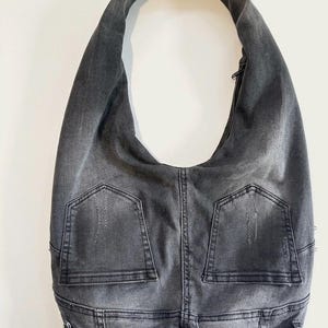 May include: A gray denim hobo bag with a single shoulder strap. The bag features two back pockets and a button closure, giving it a casual, repurposed look. The bag is made from what appears to be recycled denim.