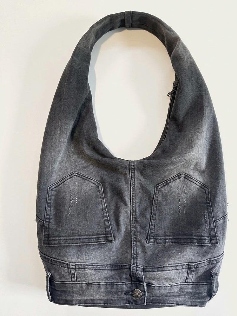 May include: A gray denim hobo bag with a single shoulder strap. The bag features two back pockets and a button closure, giving it a casual, repurposed look. The bag is made from what appears to be recycled denim.