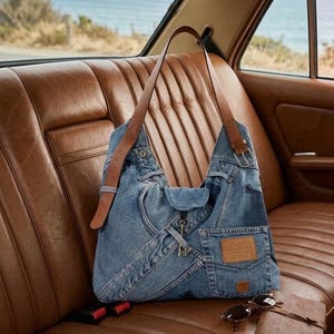 May include: A blue denim shoulder bag with brown leather straps and details. The bag has a pocket with a leather patch and a key charm. Sunglasses and a small notebook are on a brown leather car seat.