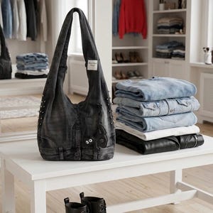 May include: A dark gray denim tote bag with silver studs and decorative stitching sits on a white table. A stack of denim jackets and jeans are next to the bag. Black ankle boots with laces are in front of the table.