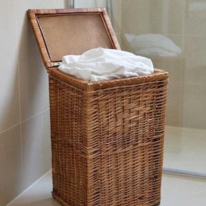 Puede incluir: Una cesta de lavandería de mimbre marrón con una tapa con bisagras, parcialmente abierta. La cesta está llena de ropa blanca doblada. La cesta es rectangular y tiene un diseño tejido. La cesta está en un baño.