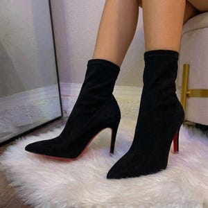 Könnte beinhalten: Ein Paar schwarze Stiefeletten mit spitzer Zehenpartie und hohem Stiletto-Absatz. Die Stiefel haben eine weiche, matte Oberfläche und eine markante rote Sohle, die unter dem Absatz sichtbar ist und bis zur Wadenmitte reicht.