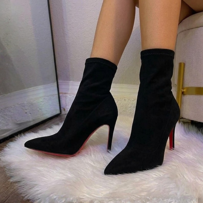 May include: A pair of black ankle boots with a pointed toe and a high stiletto heel. The boots feature a soft, matte finish and a distinct red sole visible on the bottom of the heel, extending up the shaft to mid-calf height.