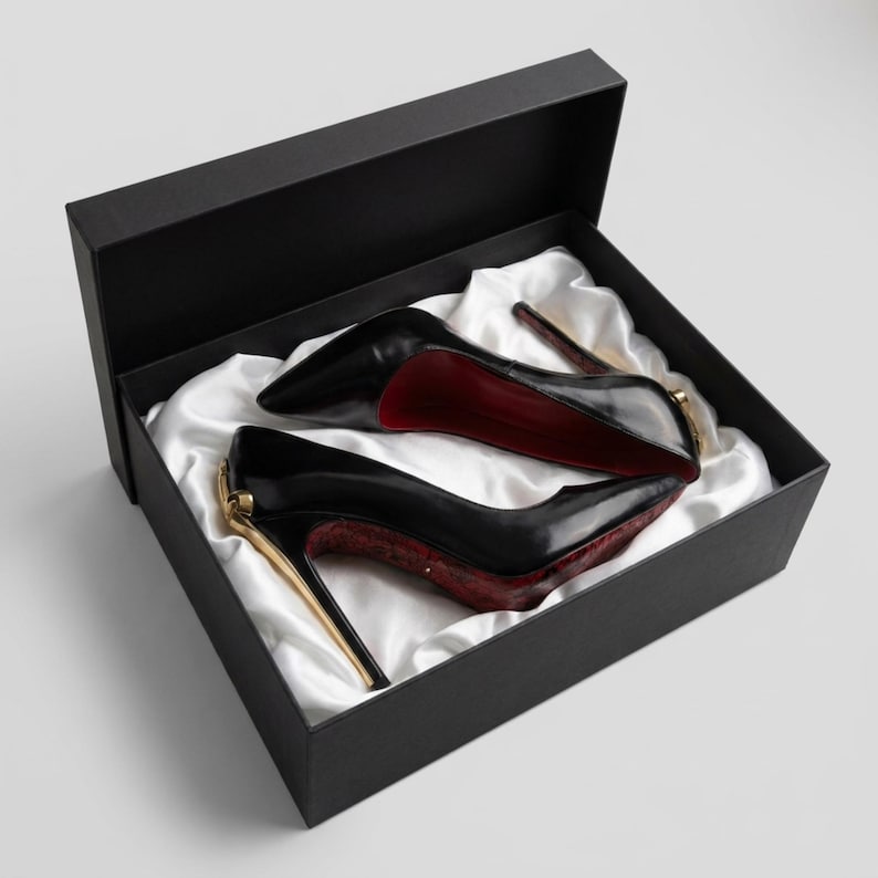 May include: A pair of black high-heeled pumps with pointed toes, gold-toned metal accents at the heels, and red interior soles. The shoes are presented inside a black rectangular box lined with white satin fabric, showcasing a sleek design.