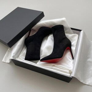 May include: A pair of black suede ankle boots with pointed toes and thin high heels featuring a signature bright red sole. The boots are presented inside a black rectangular cardboard box lined with white satin fabric and a matching lid.