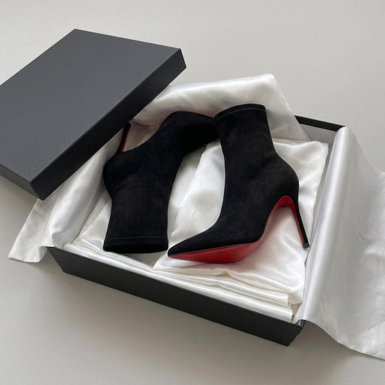 May include: A pair of black suede ankle boots with pointed toes and thin high heels featuring a signature bright red sole. The boots are presented inside a black rectangular cardboard box lined with white satin fabric and a matching lid.