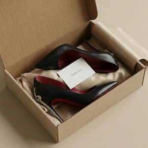May include: A pair of black high-heeled shoes with red soles and a pointed toe, presented in a cardboard box with a "Thank You" card. The shoes have a silver accent on the heel.