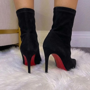 May include: A pair of black ankle boots featuring a soft, suede-like texture, a pointed toe, and a high stiletto heel. The soles of the boots are a vibrant, solid red color, providing a distinct contrast against the black upper material.