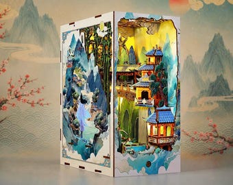 May include: A rectangular wooden book nook insert featuring a multi-layered 3D diorama of a mountain landscape with traditional architecture, blue-roofed pavilions, a bridge, and a figure in flowing robes, illuminated by warm internal LED lighting.