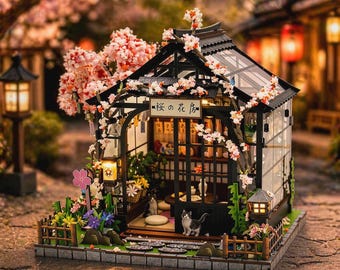 May include: A detailed miniature Japanese house with a black roof and glass walls, adorned with pink cherry blossoms. The scene includes miniature cats, flowers, and lanterns. The house has a small sign with Japanese text. The overall design is intricate and charming.