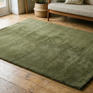 May include: A large, rectangular olive green rug with a plush, textured surface. The rug is placed on a wooden floor, and the room has a neutral color palette with a sofa and small table.