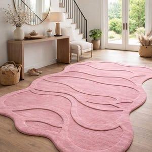 Op de afbeelding: Roze, abstract gevormd vloerkleed met een golvend patroon. Het kleed, gemaakt van zacht materiaal, ligt op een houten vloer. Het ontwerp heeft verhoogde, gebogen lijnen die textuur toevoegen. Afmetingen: ongeveer 200 cm x 300 cm.