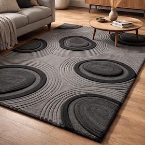 May include: A gray area rug with a circular design. The rug features concentric circles in varying shades of gray and black, creating a textured, dimensional effect. The rug is placed on a hardwood floor.