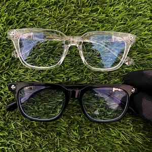 May include: Two pairs of eyeglasses with rectangular frames, one in clear transparent acetate and one in solid black acetate. Both feature silver-toned metallic accents on the temples and hinges, with the Chrome Hearts logo visible on the frames.