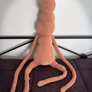 May include: A peach-coloured, handmade crochet plush toy with a segmented body, long limbs, and two black pom-pom antennae. The soft, textured toy is designed to be a whimsical creature.