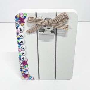 May include: A white wooden photo holder with a decorative beaded border in pink, blue, and green. The holder has a twine bow and a metal clip. The word "RACHA" is spelled out with white letter beads.