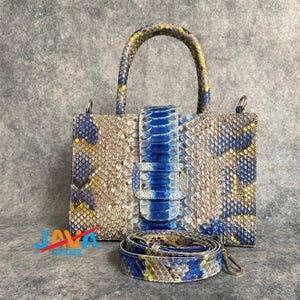 Puede incluir: Bolso de mano rectangular con asa superior y bandolera extraíble. El bolso presenta un estampado de piel de serpiente en tonos azules, amarillos y beige. Una correa azul con hebilla plateada asegura la parte delantera. El bolso tiene herrajes plateados.