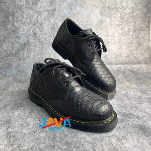 May include: Black leather shoes with a textured snakeskin pattern. The shoes have black laces, eyelets, and soles with yellow stitching. The shoes are displayed on a gray surface.