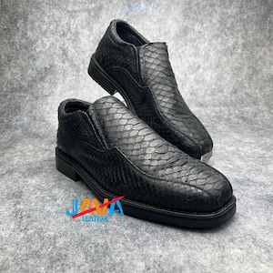 May include: A pair of black leather slip-on shoes with a textured snakeskin pattern. The shoes have a low-profile design with a rounded toe and a thick sole. The image also shows the logo "JAVA LEATHER".