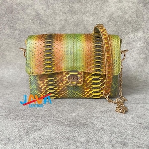 Puede incluir: Un pequeño bolso rectangular con un colorido estampado de piel de serpiente en tonos verdes, amarillos y naranjas. El bolso tiene una correa de cadena dorada y un cierre de broche. El texto "JAVA LEATHER" es visible.